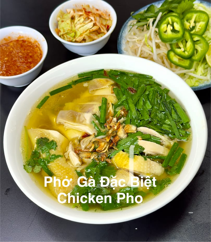 A bowl of chicken pho with broth, chicken, green onions, and cilantro, with side dishes.